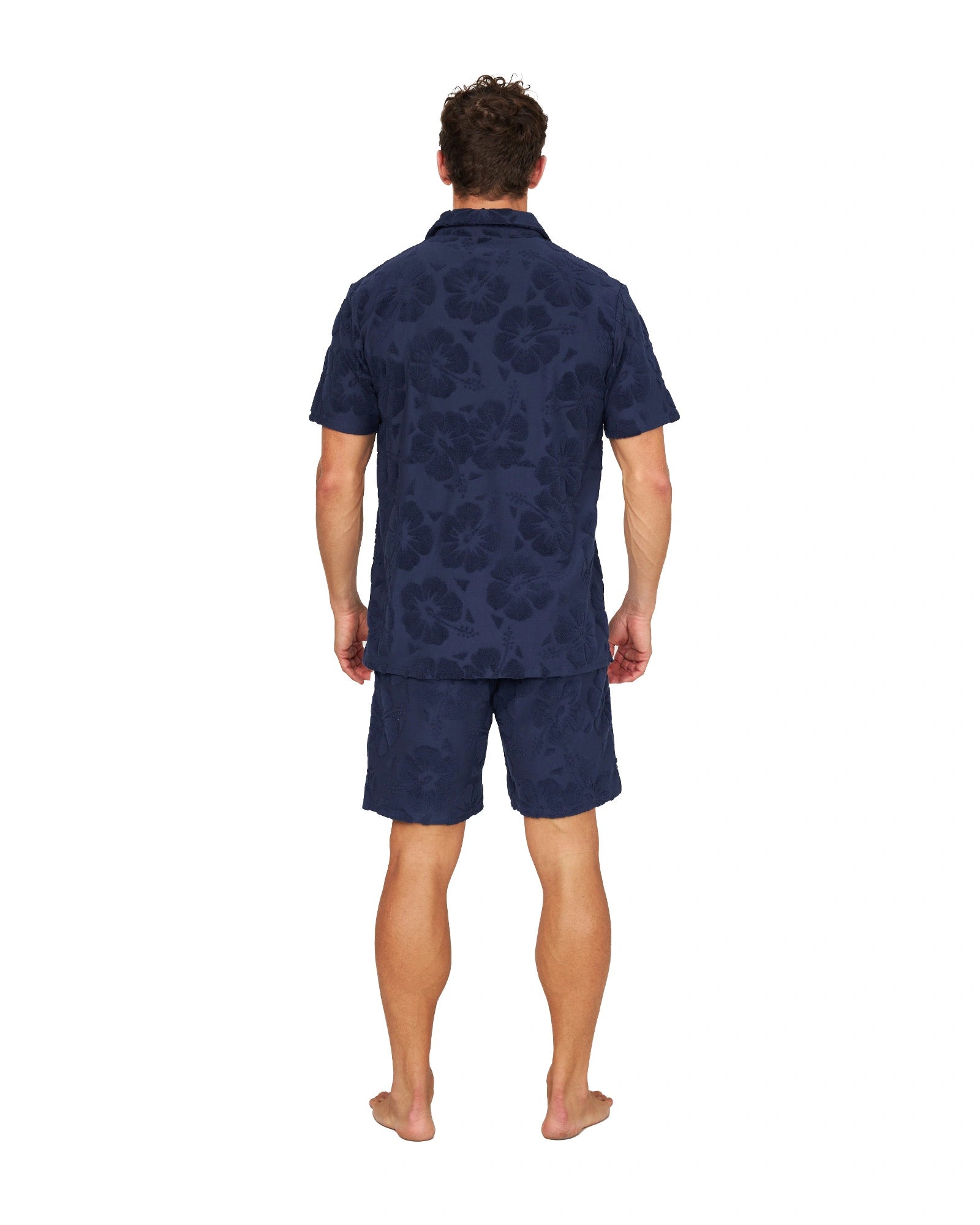 A full body back view of a male model wearing the Okanui Terry Jacquard Shirt in Hibiscus Navy and terno shorts.