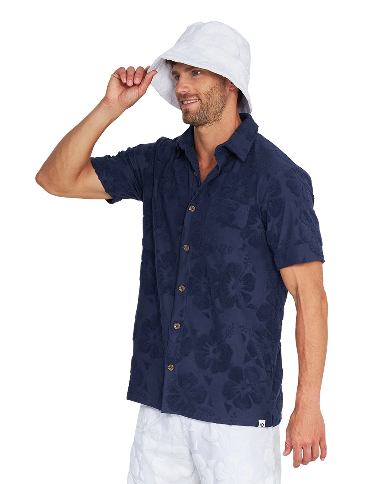 A male model wearing the Okanui Terry Jacquard Shirt in Hibiscus Navy, a white bucket hat and white shorts.