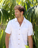 A male model with a small palm tree in the background wearing the Okanui Starboard Short Sleeve Shirt in Hibiscus white.