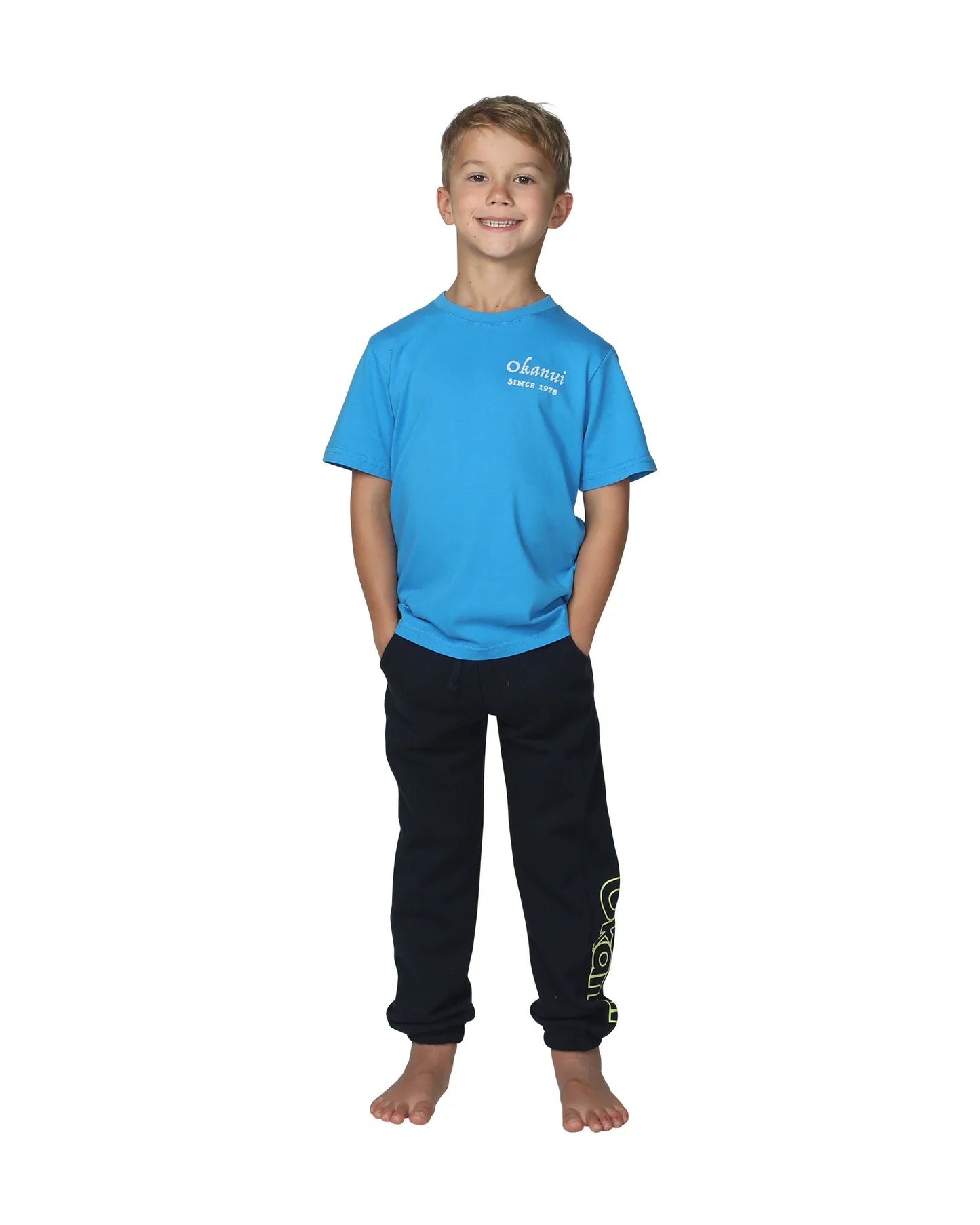 Full body front view of a little boy wearing the Okanui Winter Keyline trackies for Kids in Navy color paired with a washed blue color Okanui T-shirt