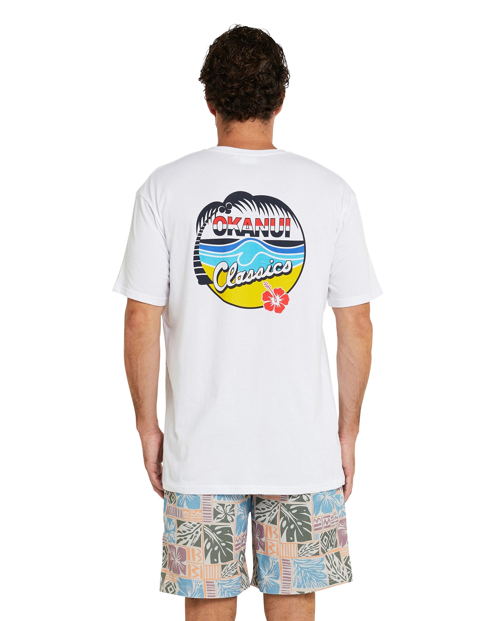 Back view of a model wearing the Okanui Classic Badge T-shirt showing the back print of the t-shirt with the Okanui logo and the word "classics" inside a beach cartoon..