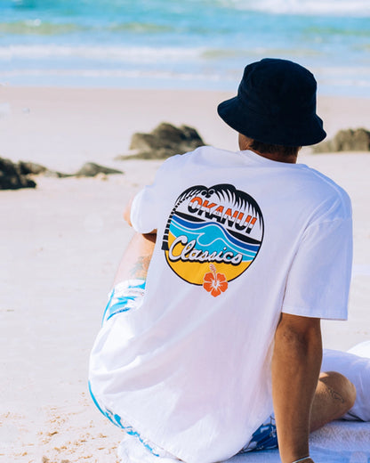 A male model sitting on the beach looking far away into the horizon wearing the Okanui Classic Badge T-shirt for men in white color showing the back print of the t-shirt.