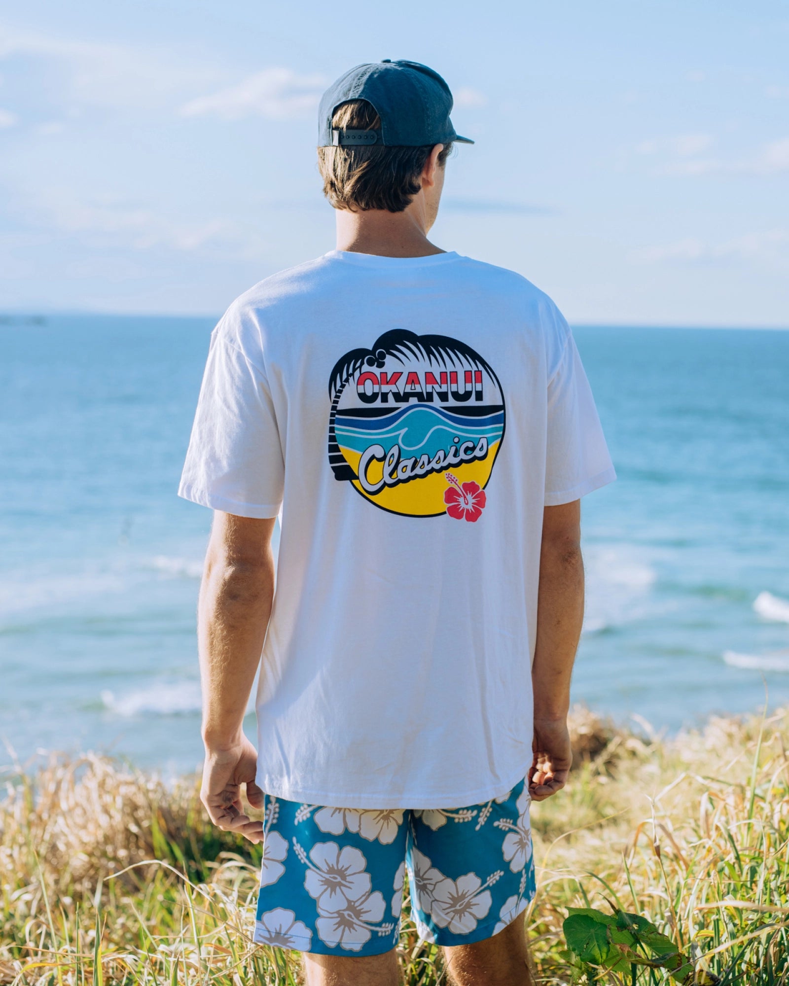 A back view model in a beach looking far away wearing a baseball cap, a hibiscus print pattern short and the Okanui Classic Badge T-shirt.