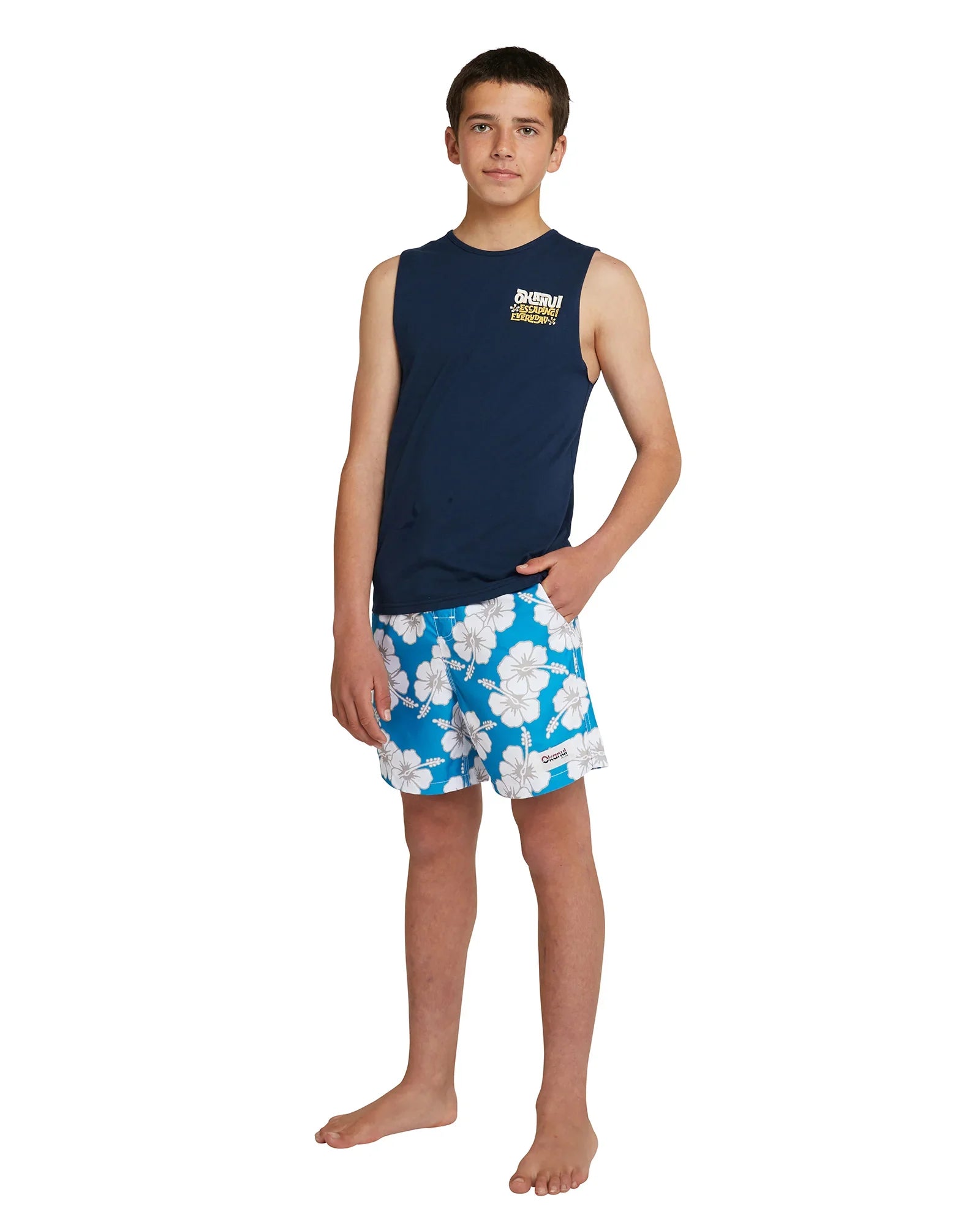 Full body front view of a boy model wearing the Okanui Boys Swim Short in Storm Blue paired with a navy tank top