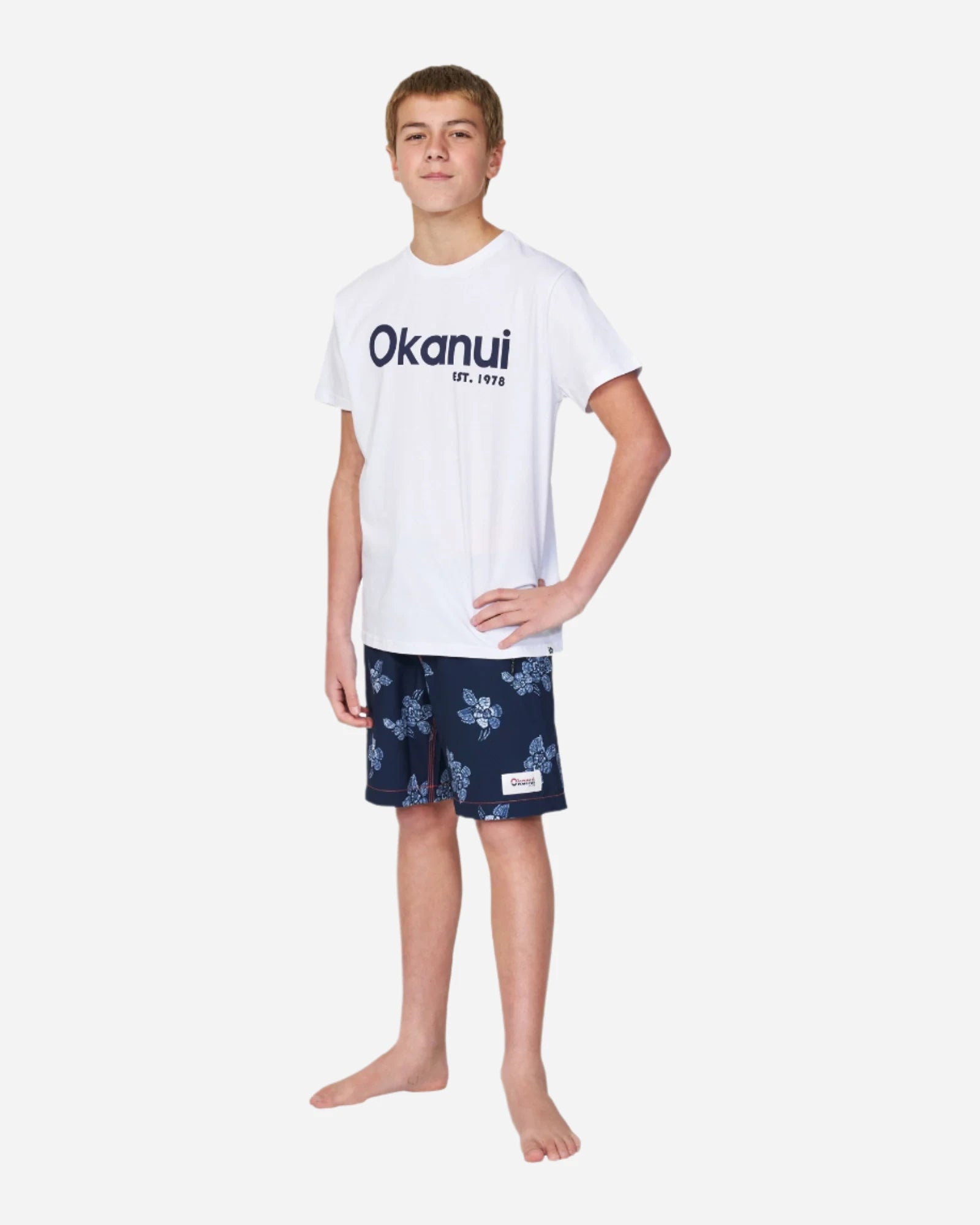 Full body front view of a boy with his hand in the hip pose wearing a white tshirt paired with Okanui boardrider board shorts for kids in White Navy