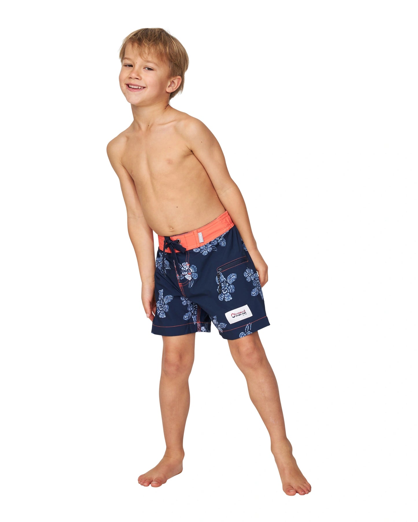 A topless little boy leaning on the side wearing the Okanui boardrider board shorts for kids in White Navy