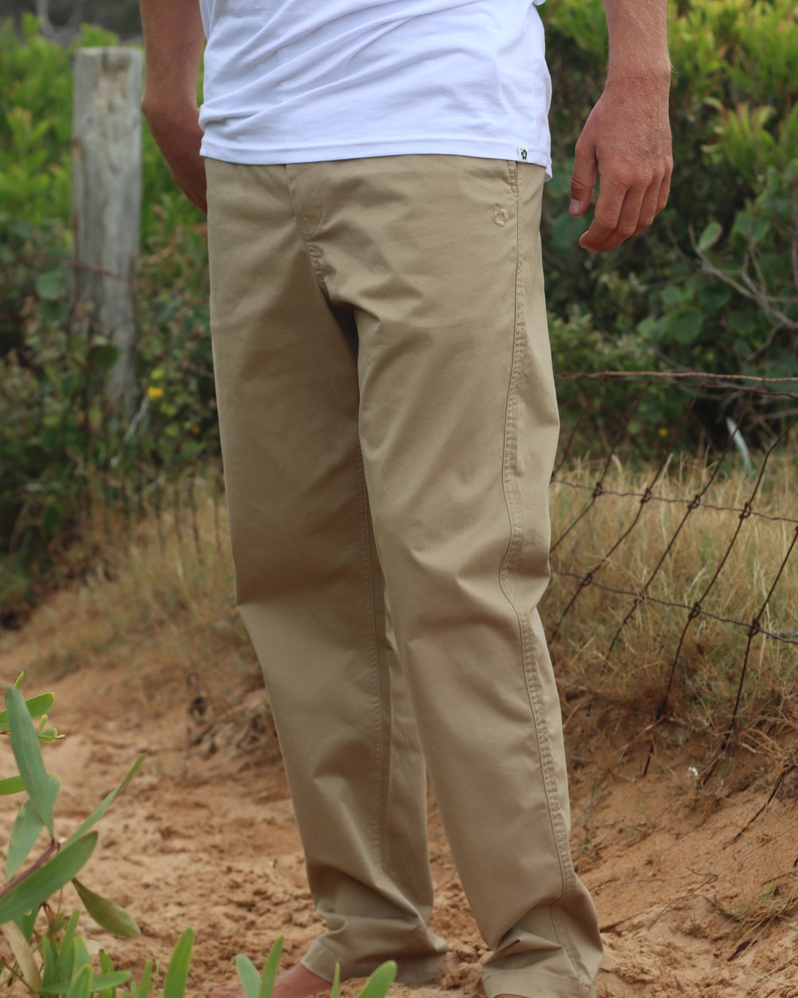 The Okanui Men's Chino Pants worn by a model outdoor with bushes and a wire fence on the background.