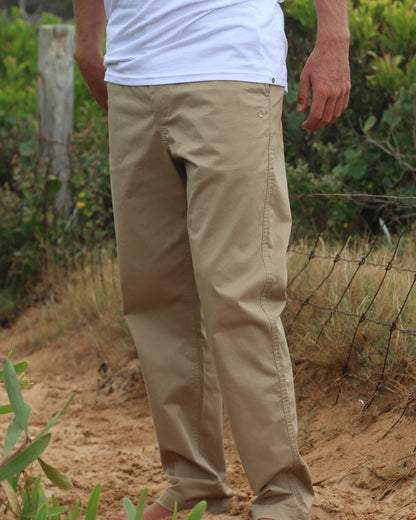 The Okanui Men's Chino Pants worn by a model outdoor with bushes and a wire fence on the background.