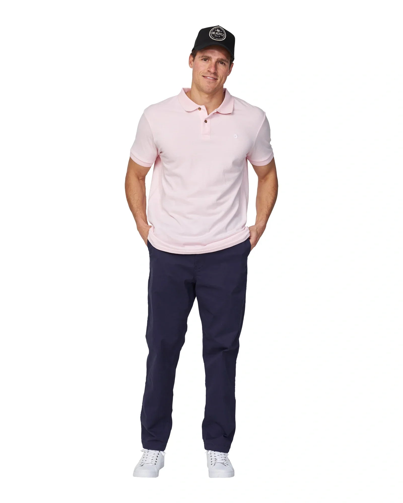 A male model wearing a black trucker hat, navy pants, white shoes and the Okanui Classic Polo shirt in pale pink colour with his hands on his pocket.