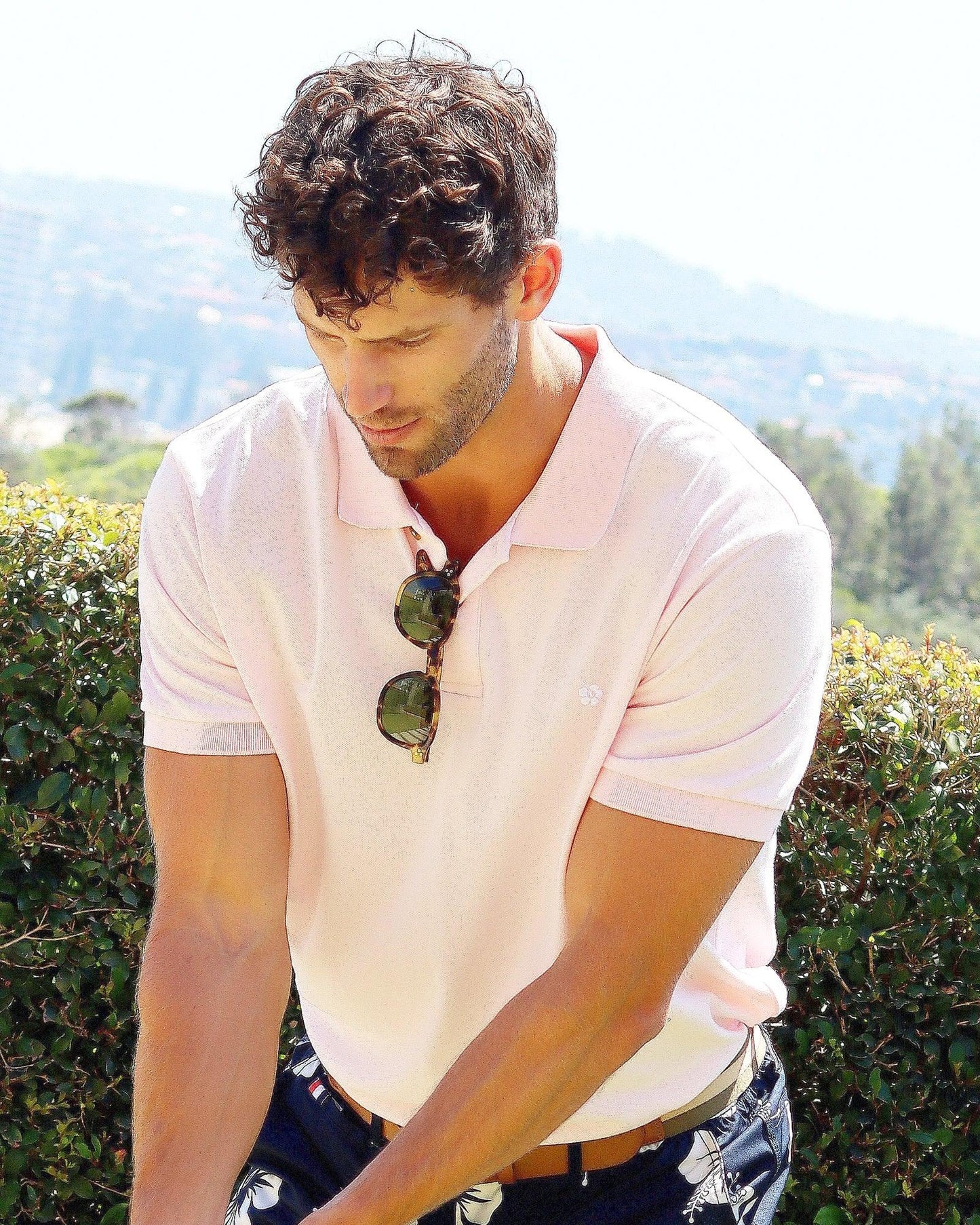 A male model wearing a pink Okanui Classic Polo shirt prepare to strike a golf ball with his glasses hanging on the neck button.