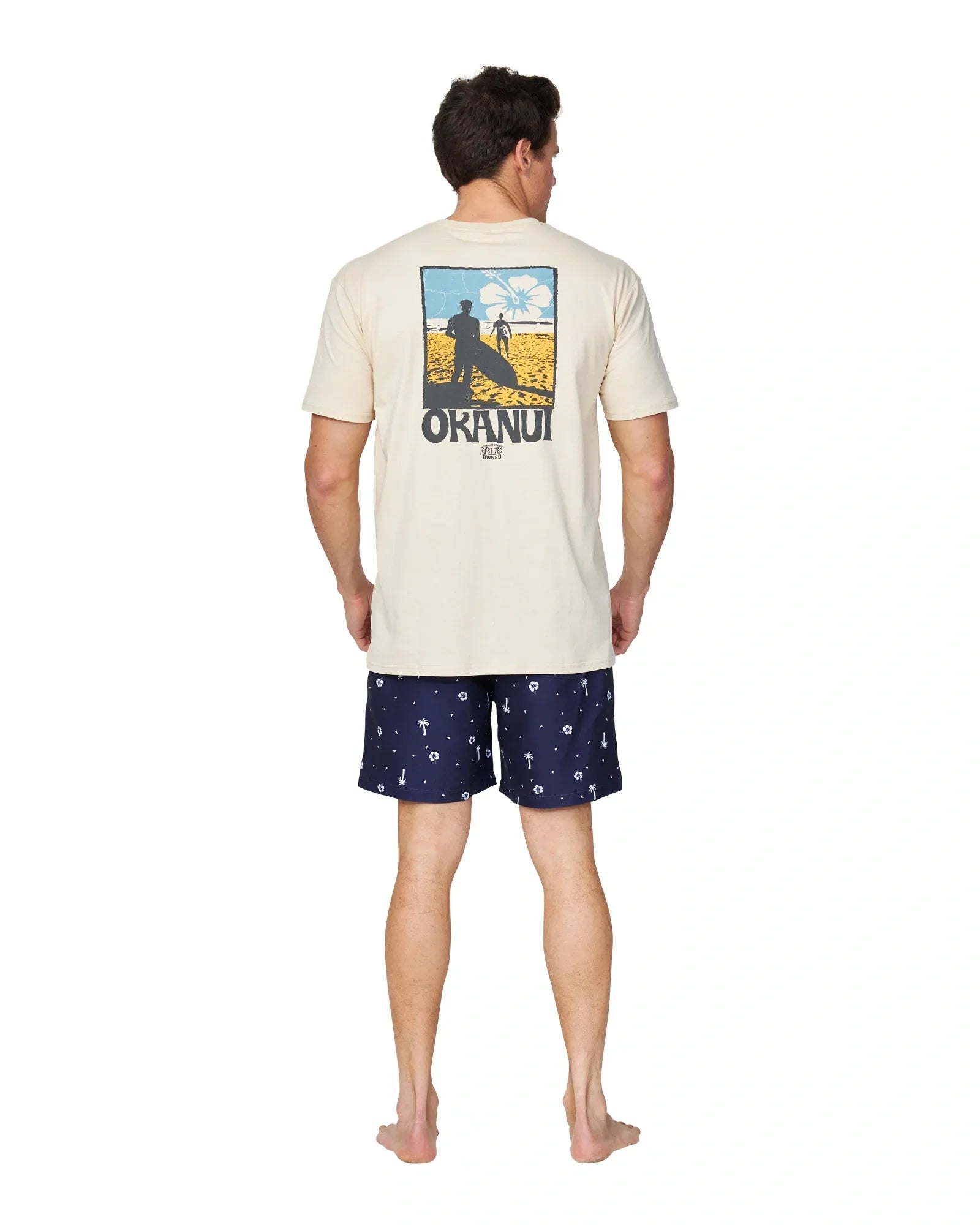 A back view pose of a model wearing a birch color t-shirt with a print on the back and the Okanui Icons Swimshorts in navy colour.