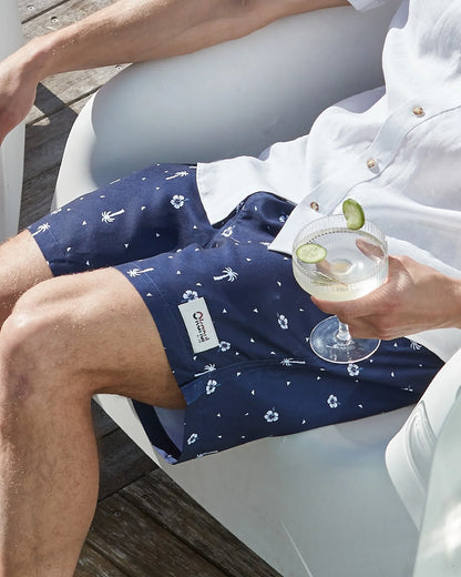 A male model sitting on a white couch holding a cocktail wearing a white button up shirt and the Okanui Icons Swimshorts in navy color.