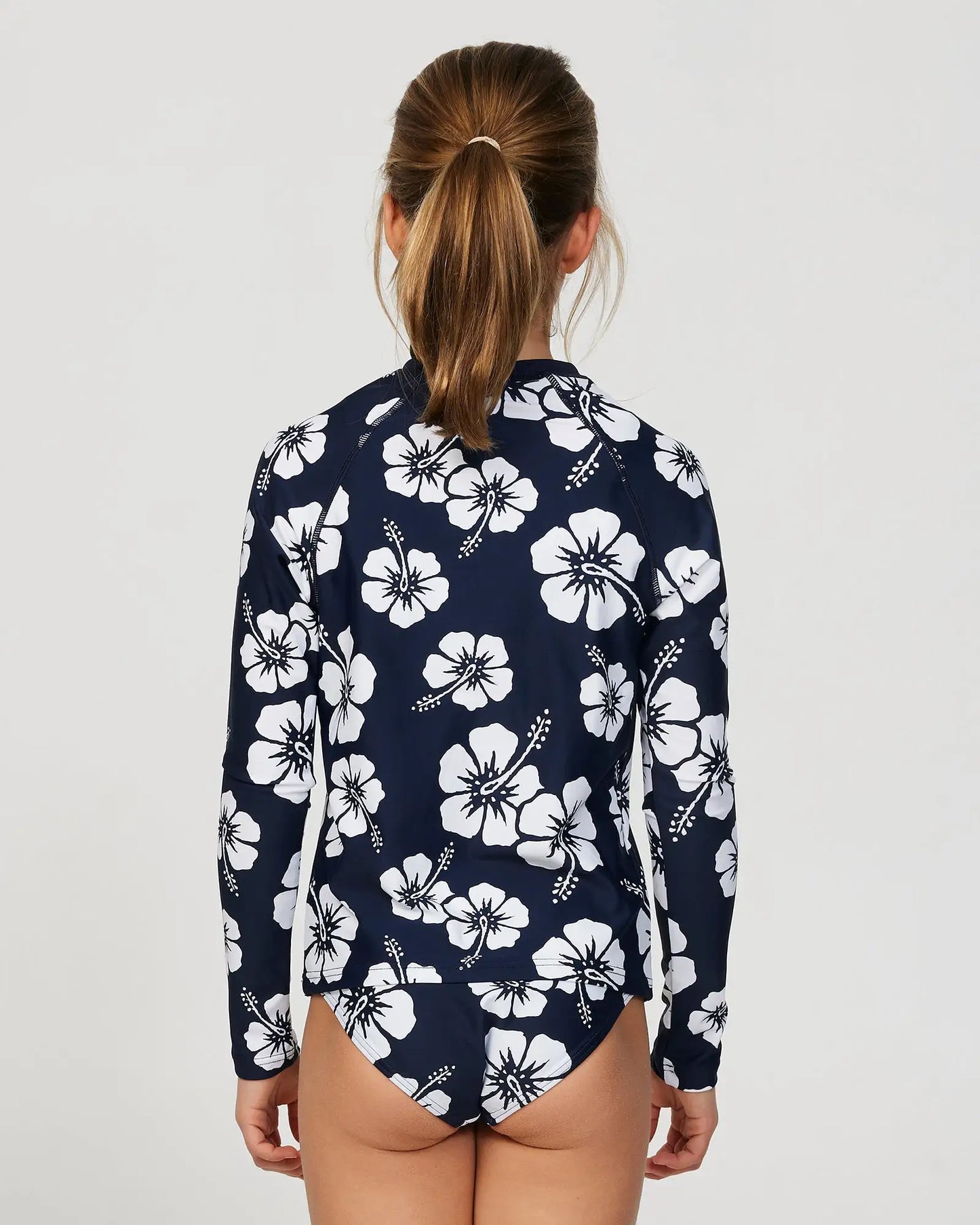 Closeup back view of a little girl model wearing the Okanui Longsleeve swimsuit for Girls in Hibiscus Navy