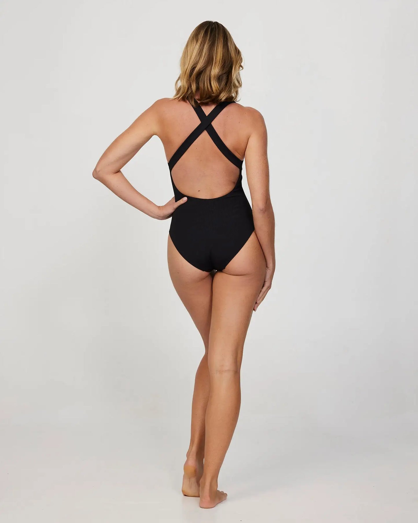 Full body back view of a female model wearing the Okanui Ribbed Cross Back One Piece Bikini in Black