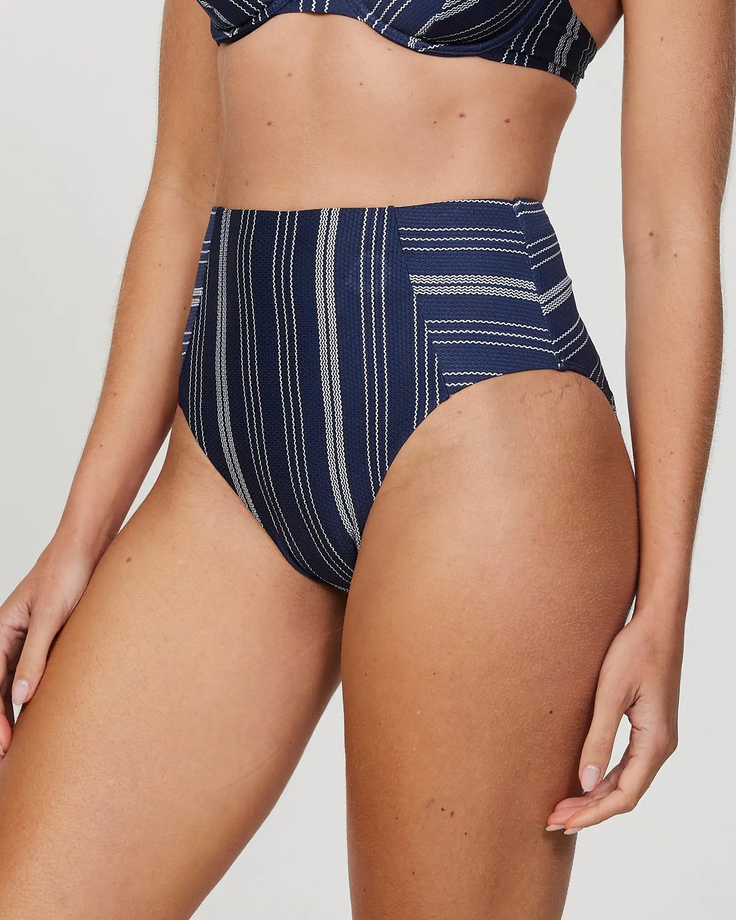 Front-left view of a female model wearing the Okanui Horizon High waist Bikini Bottom in Navy Stripe