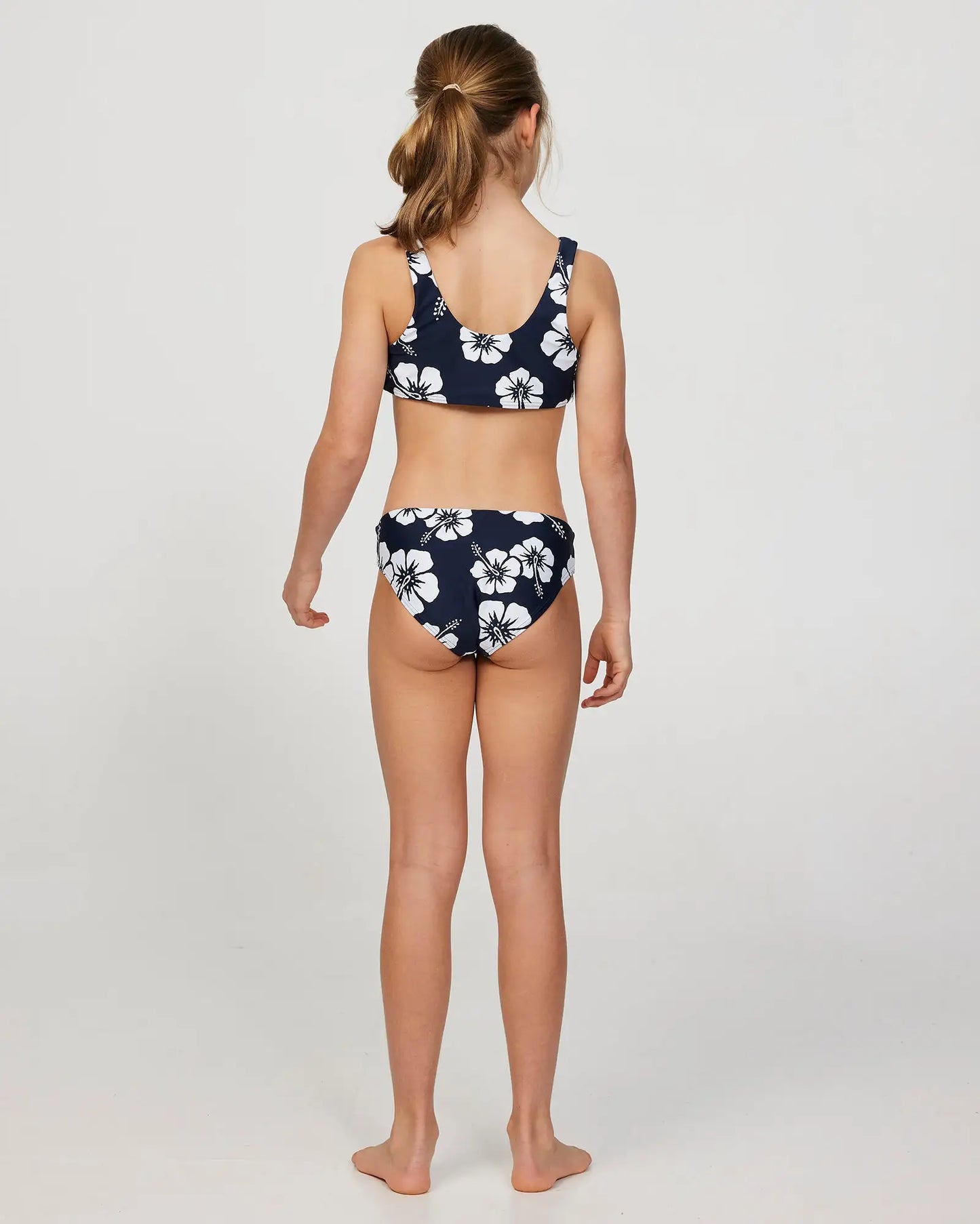 Full body back view of a little girl model wearing the Okanui Crop Bikini Set in Hibiscus Navy