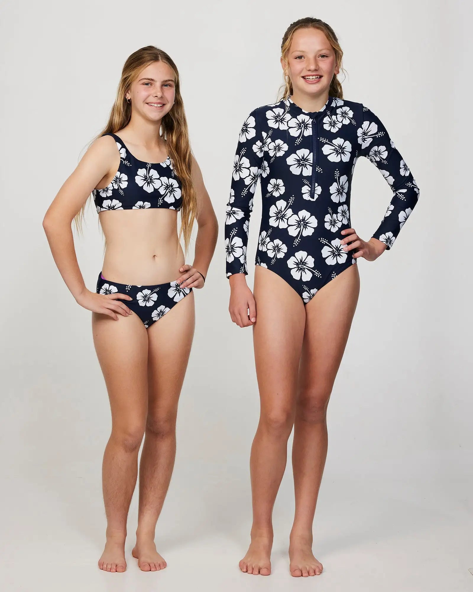 Two girl models wearing the Okanui Crop Bikini Set and long sleeve one piece swimsuit in Hibiscus Navy