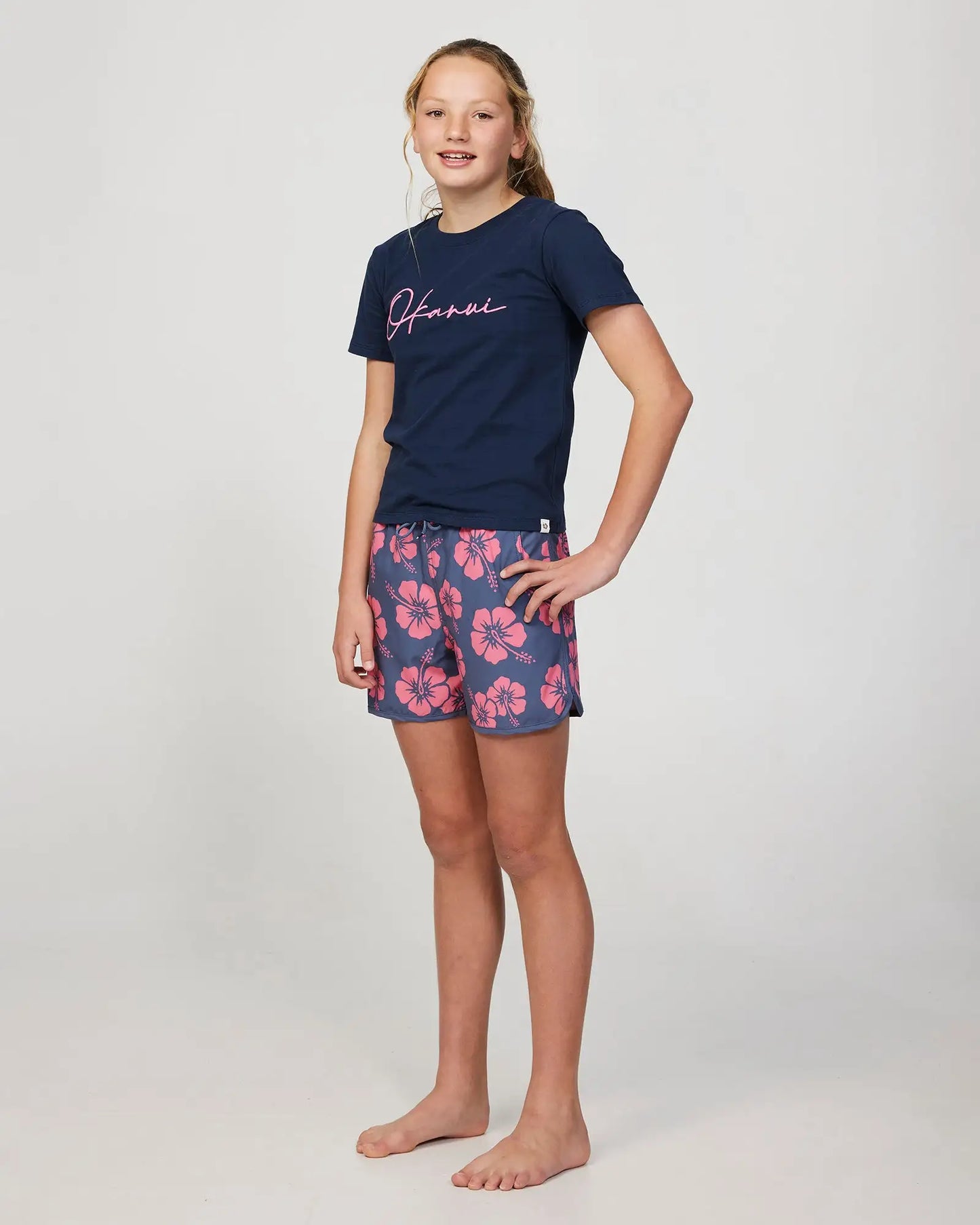 Full body front-left view of a girl model wearing the Okanui swim short for girls in Hibiscus Iron Pink