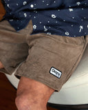 A close up lower front view of a male model sitting on a white couch wearing a navy button up shirt and the Okanui Big Iron Cord Walk Short featuring it's curdoroy cotton cloth in stone colour.