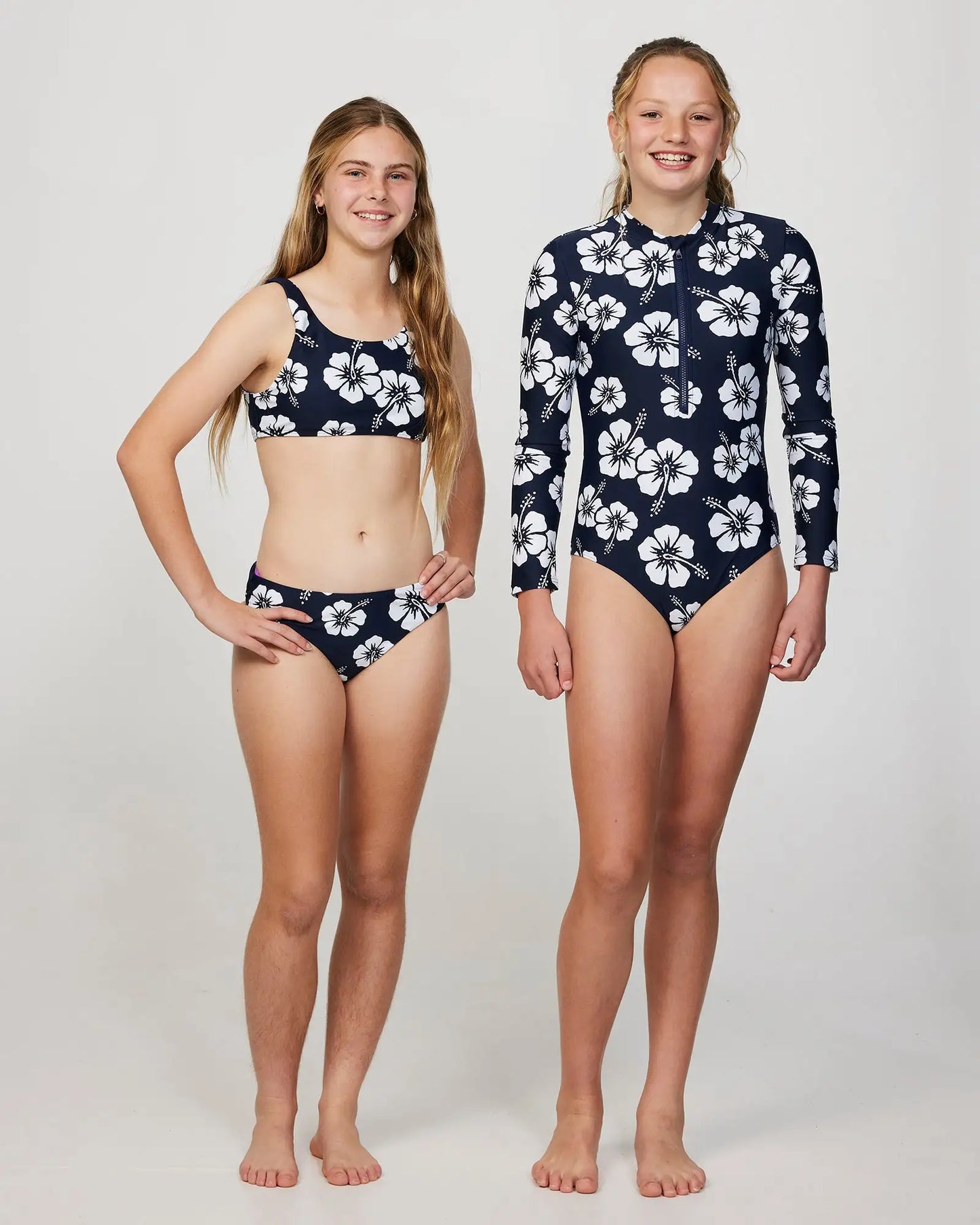 Two girl model wearing the Okanui long sleeve one piece swimsuit for girl in Hibiscus Navy and swim crop top on the left girl