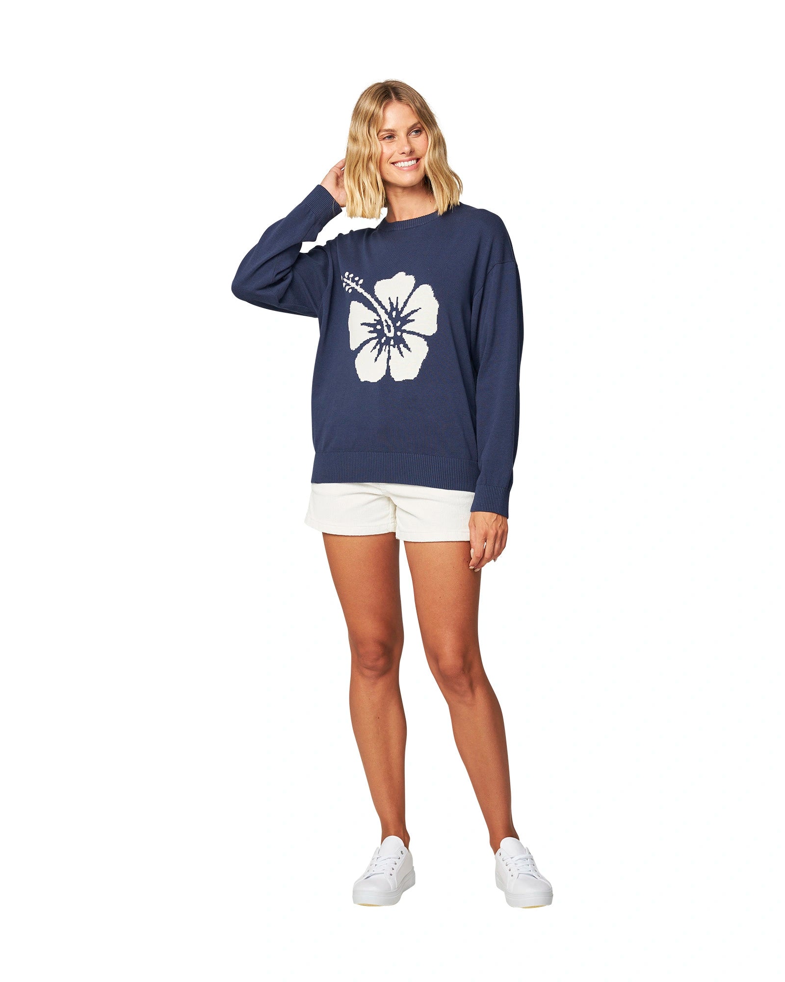 Full body front view of a female mode with her one hand in her hair, wearing the Okanui waffle knit jumper in navy color paired with white shorts