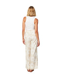 Back view of a female model wearing a white tank top paired with the Okanui OG Paradise wide leg beach pants in Hibiscus Natural color