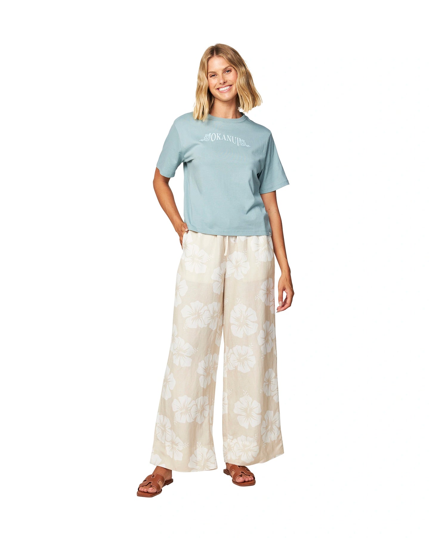 Full body front view of a female model wearing a blue shirt paired with the Okanui OG Paradise wide leg beach pants in Hibiscus Natural color