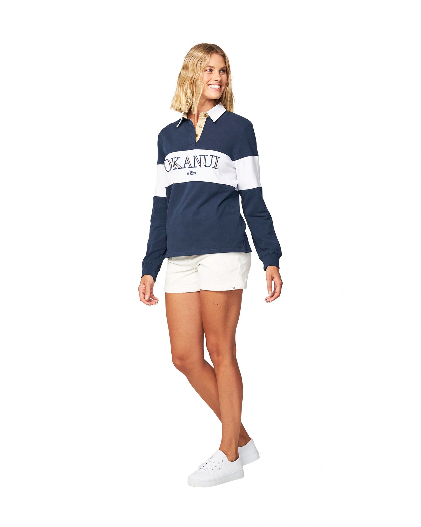 A female model wearing the Okanui Heritage rugby top jumpers in navy/white color paired with white shorts