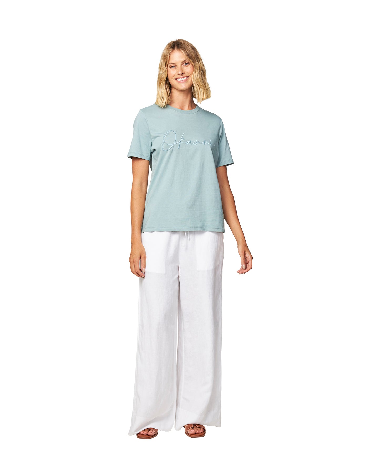 Full body front view of a female model wearing the Okanui Signature short sleeves T-shirt in Sage color paired with white trousers