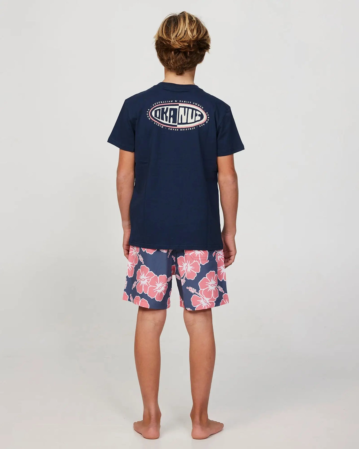 Full body back view of a boy model wearing the Okanui Way Back When Stretch Swim Short for Boys in Iron Pink