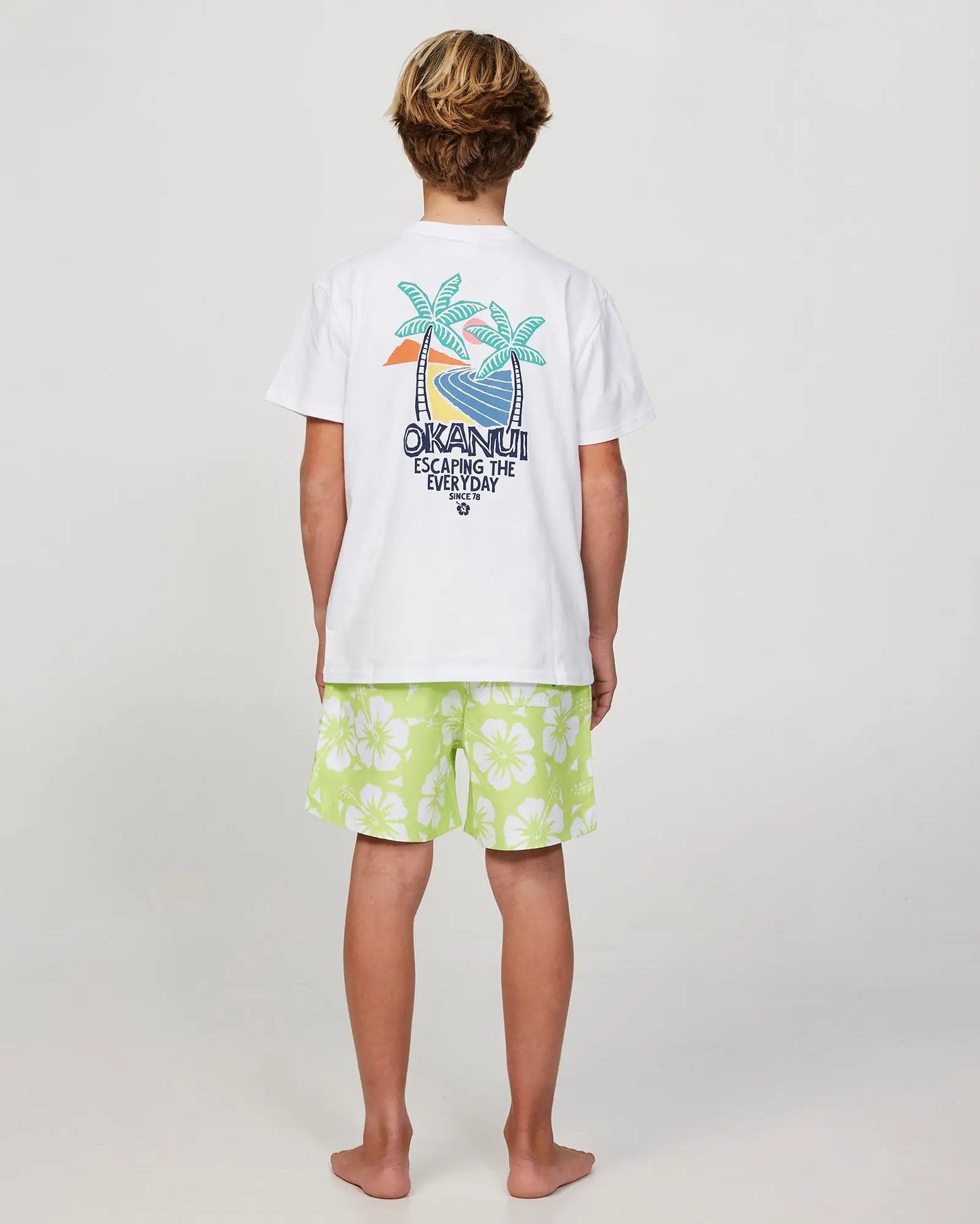 Full body back view of a boy model wearing the Okanui Swim Short for boys in Hibiscus Washed Lime