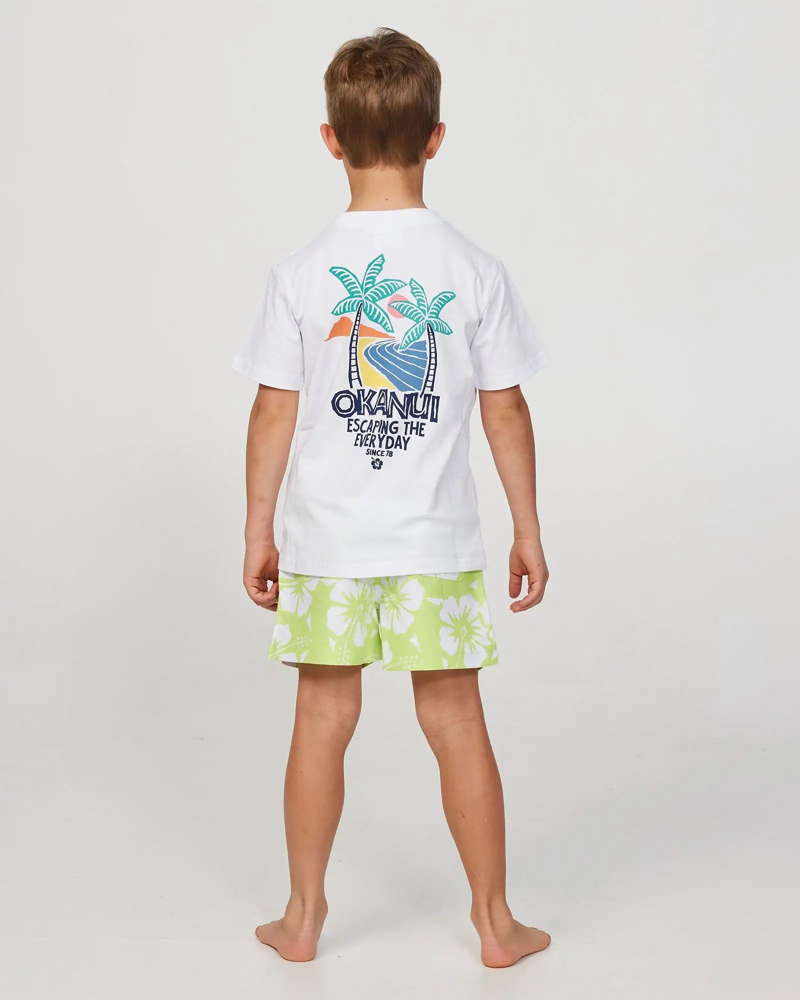 Full body back view of a little boy model wearing the Okanui Swim Short for boys in Hibiscus Washed Lime