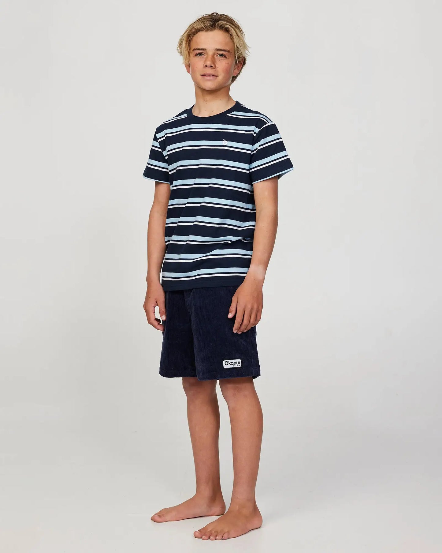 Full body front view of a boy model wearing the Okanui Staple Stripe T-shirt for boys in Navy Multi