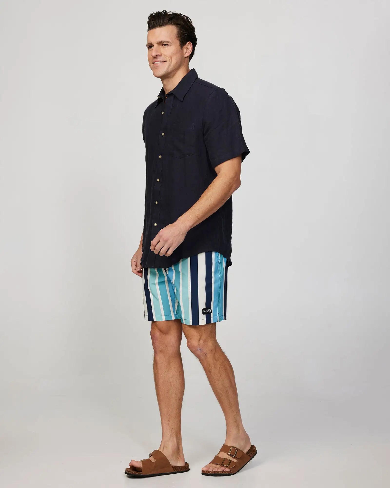 Full body Left side view of a male model wearing the Okanui Mens Straight Lines Stretch Swim Short in Stone Multi paired with a black button up shirt