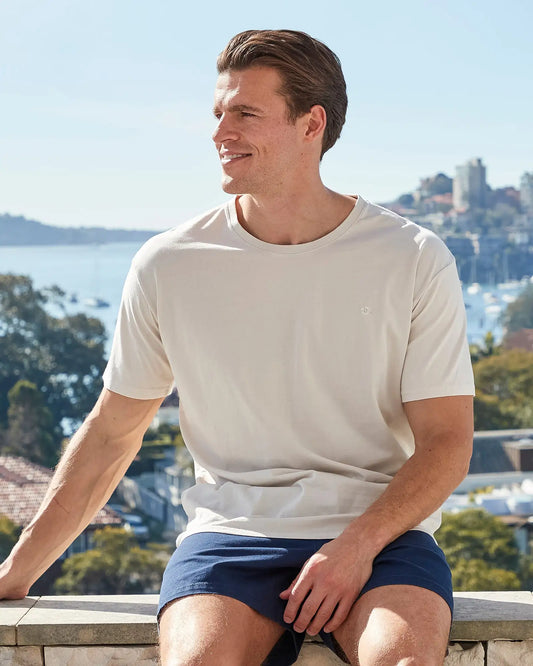 A male model sitting on a ledge wearing the Okanui Mens Staple Tee T-shirt in Stone