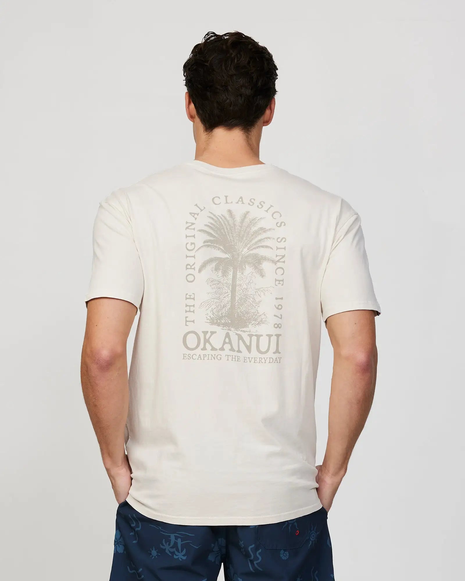 Back view of a male model wearing the Okanui Mens Barrel Tee T-shirt in Stone