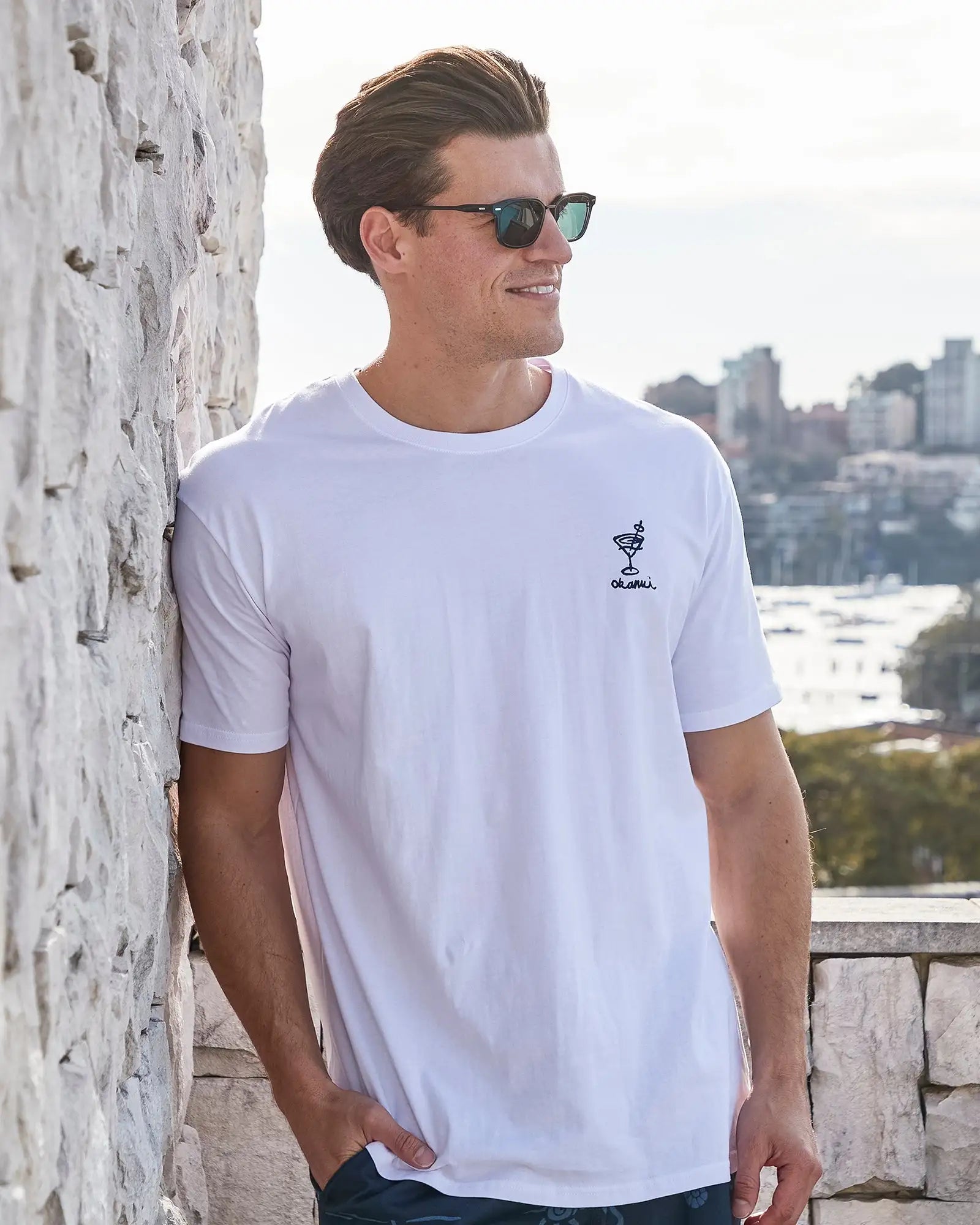 Front view of a male model wearing sunglasses leaning on the wall wearing the Okanui Cruise Tee T-shirt in White