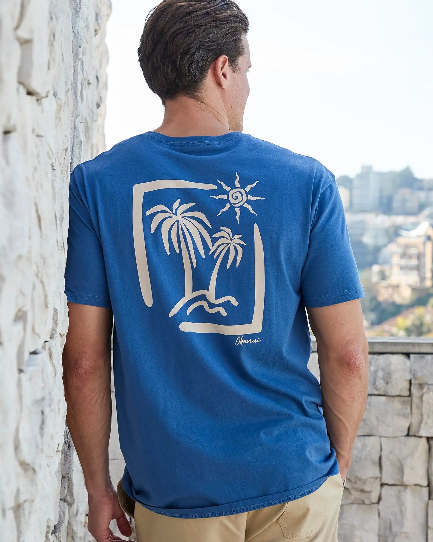 Back view of a male model leaning on the wall wearing the Okanui Mens Ripple Tee T-shirt in Washed Navy