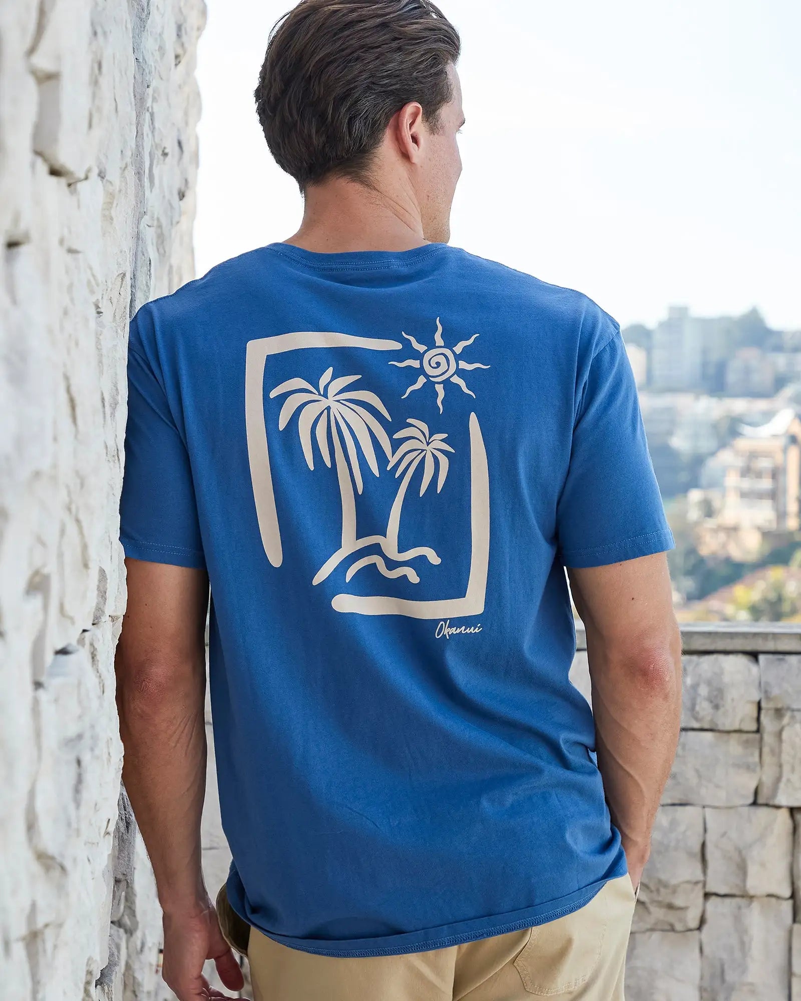 Back view of a male model leaning on the wall wearing the Okanui Mens Ripple Tee T-shirt in Washed Navy