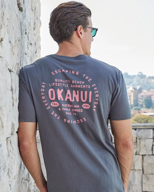 Back view of a male model leaning on a wall wearing the Okanui Mens Stampede Tee T-shirt in Washed Black