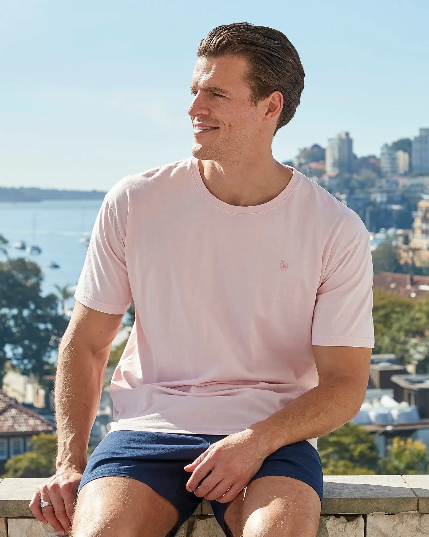 Front view of a male model sitting on a ledge wearing the Okanui Mens Staple Tee T-shirt in pink