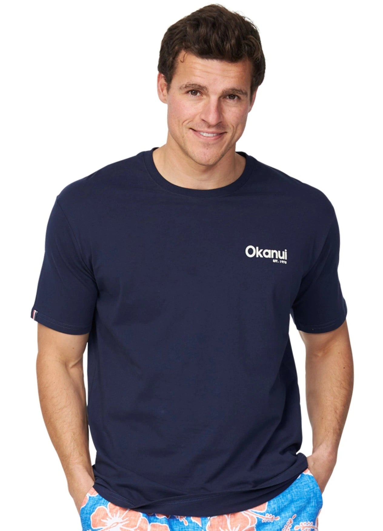 A closer front view of a male model with his hands in his pockets and wearing the Okanui Classic Badge Tee in navy color.