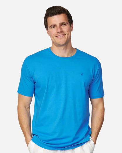 The Okanui Men's Staple Tee in washed blue color is a simple blue tshirt with a small Hibiscus logo on the left chest and it is worn by a smiling model.