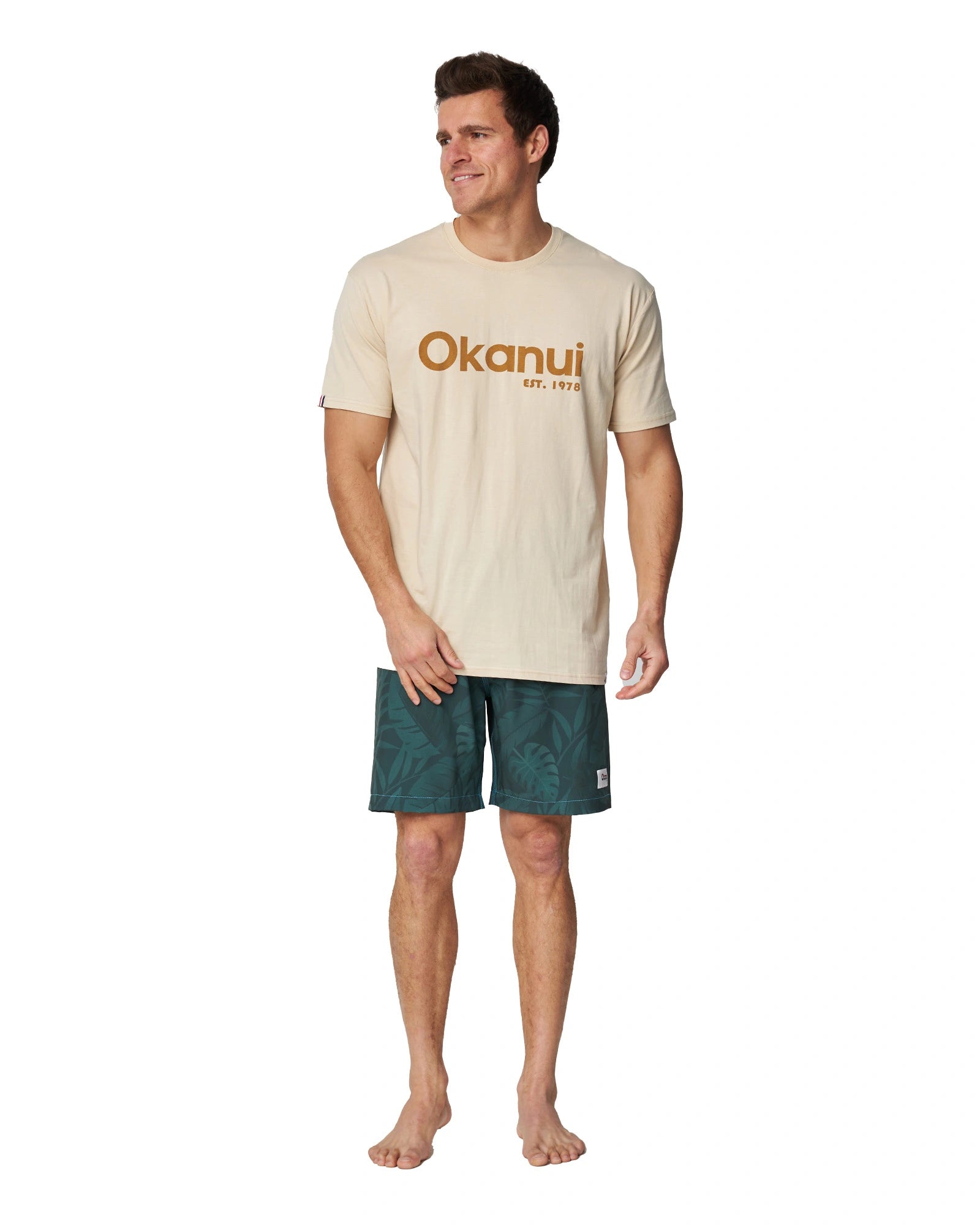 A full body front view of a male model wearing the Okanui Men's Logo Tee in birch color.