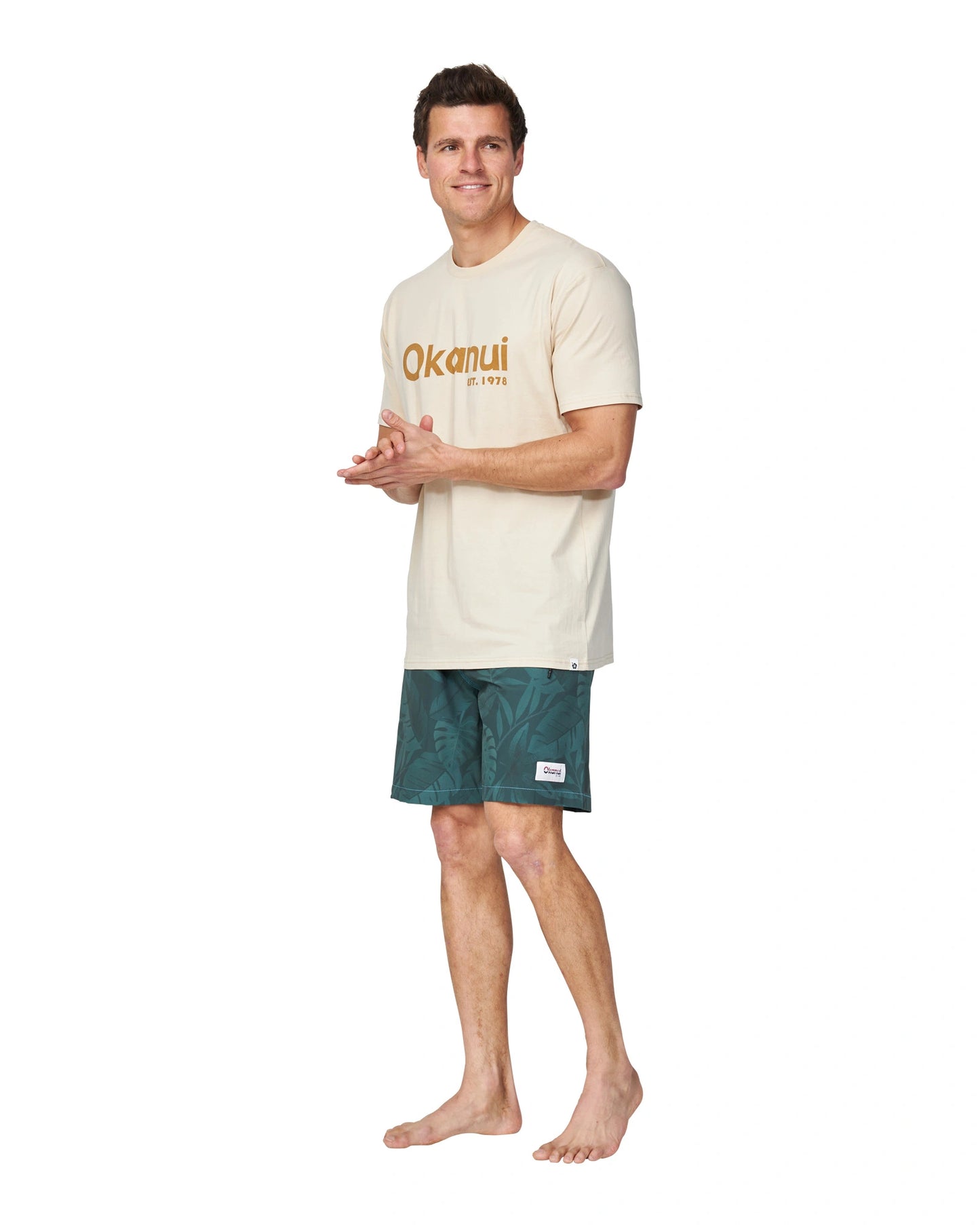 A full body angle of a male model wearing the Okanui Men's Logo Tee in birch color.