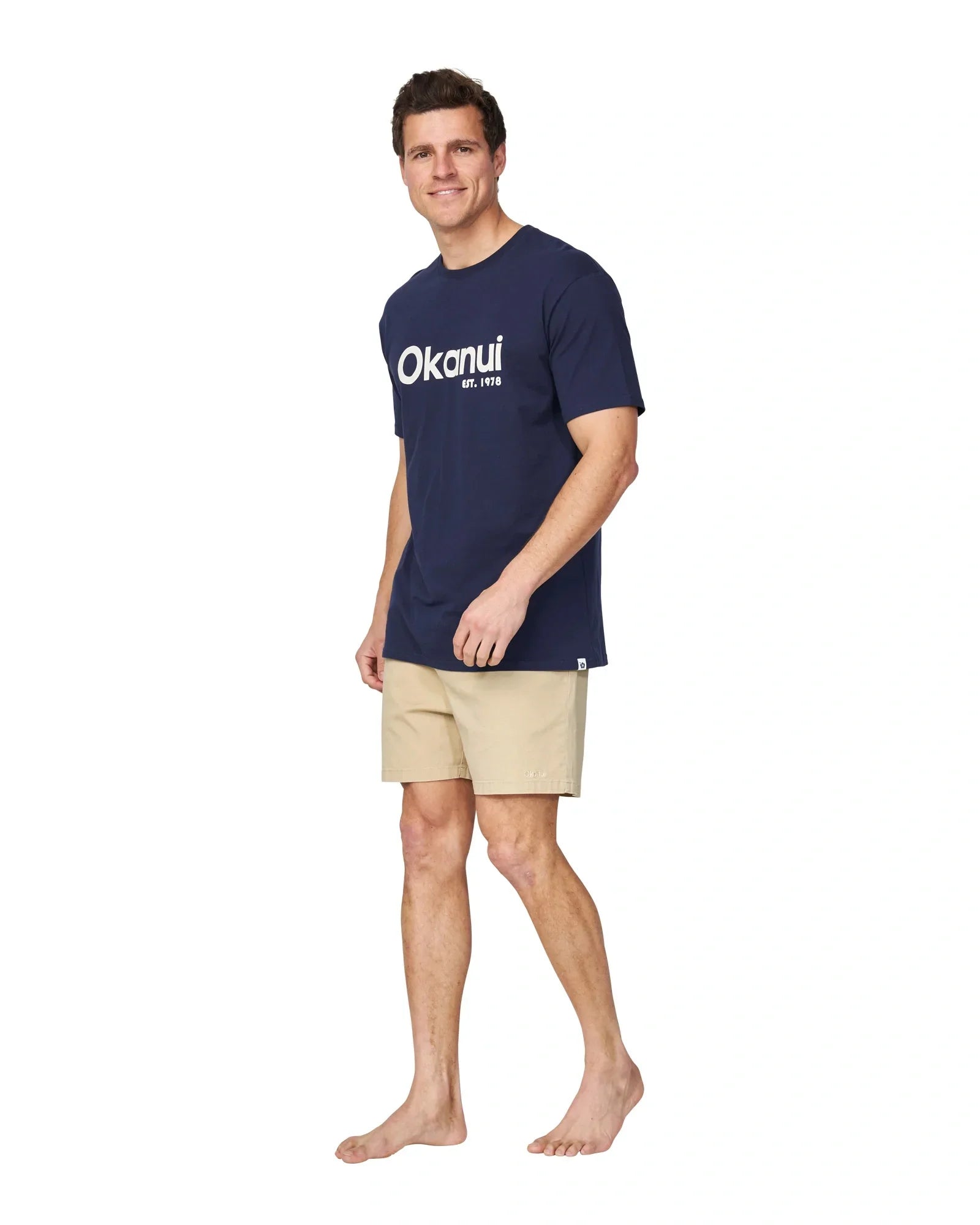 A full body slightly side view of a male model wearing the Okanui Logo Tee in black.